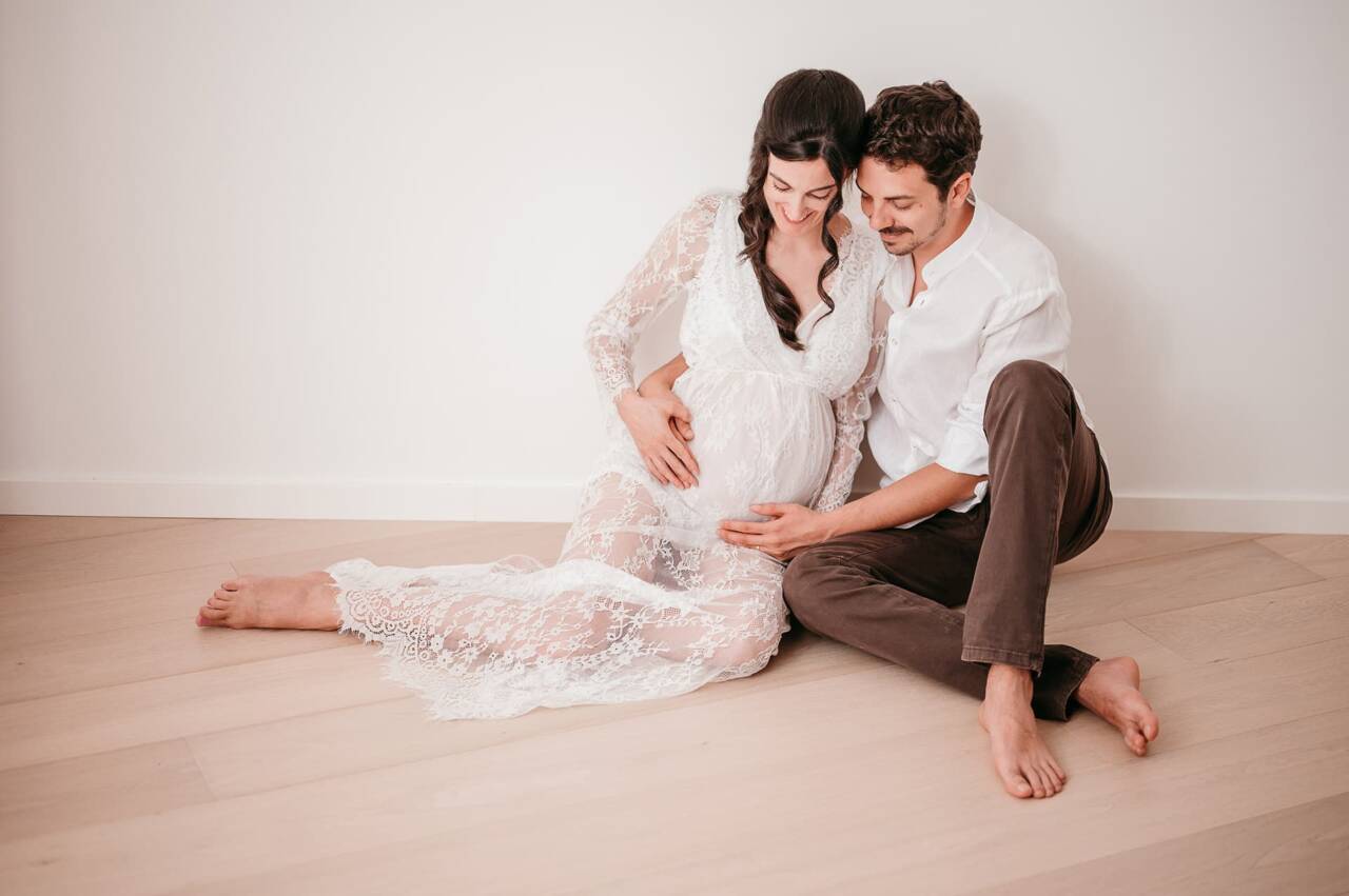 Futuri genitori fotografati al momento giusto in stile minimal con abito bianco di pizzo seduti sul pavimento durante il loro servizio fotografico di gravidanza